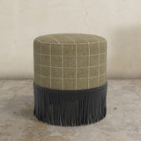 Round ottoman with plaid top and fringed bottom on a concrete floor.