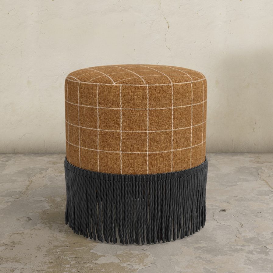 Round ottoman with plaid top and fringed bottom on a concrete floor.