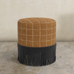 Round ottoman with plaid top and fringed bottom on a concrete floor.