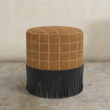 Round ottoman with plaid top and fringed bottom on a concrete floor.