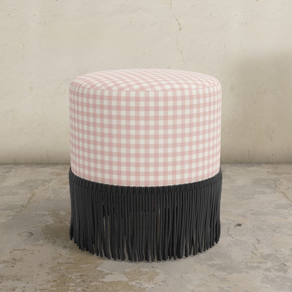 Blush pink gingham upholstered round ottoman with white cushion and fringes on a concrete floor.