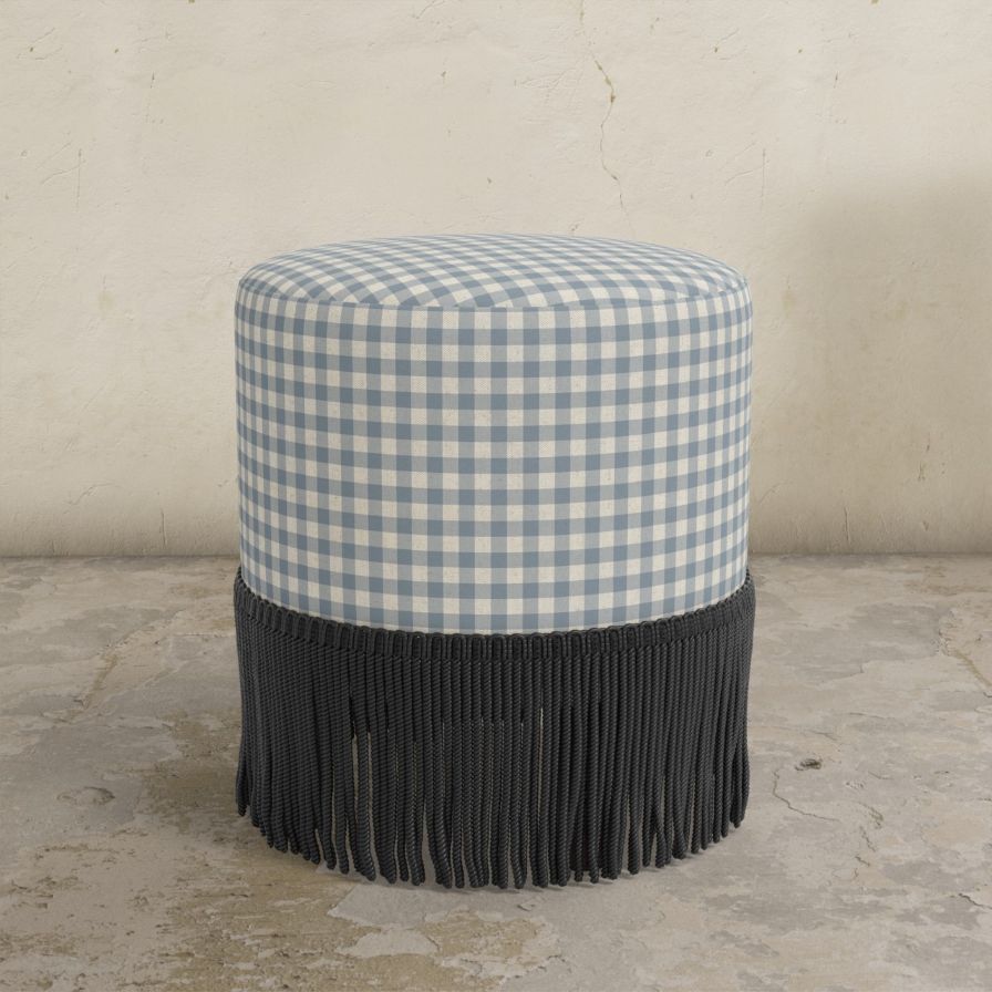 Round upholstered ottoman with blue and white gingham top and black fringed bottom on a concrete floor.