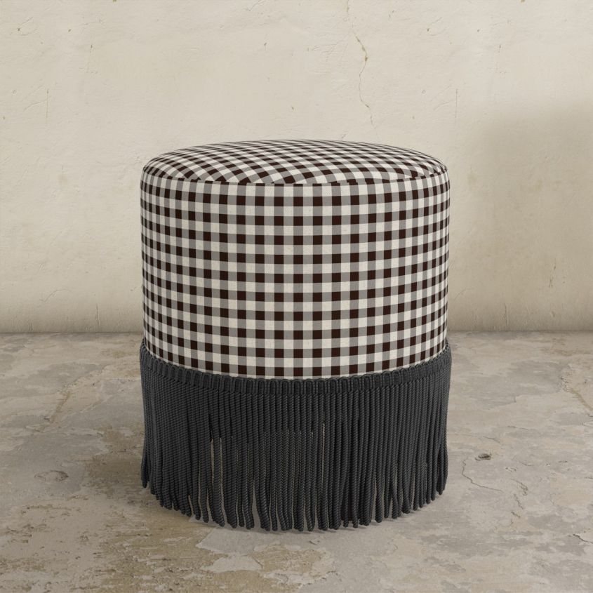 chocolate brown gingham upholstered ottoman with fringe on a concrete floor.
