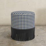 Round upholstered ottoman with gingham check and fringes on a concrete floor.