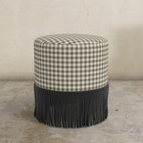 Round upholstered ottoman with fringes on a concrete floor.