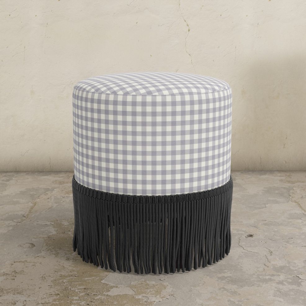 Classig gingham lilac upholstered ottoman with black fringe on a concrete floor.