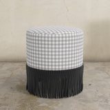 Classig gingham lilac upholstered ottoman with black fringe on a concrete floor.
