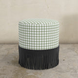 Round moss green gingham upholstered ottoman with black fringes on a concrete floor.