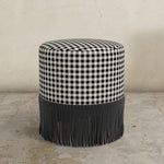 Black gingham round upholstered ottoman with fringed bottom on a concrete floor.