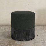 Black upholstered ottoman with fringe on a concrete floor and light gray wall background