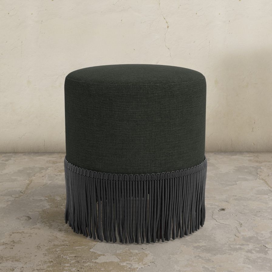 Black upholstered ottoman with fringe on a concrete floor and light gray wall background
