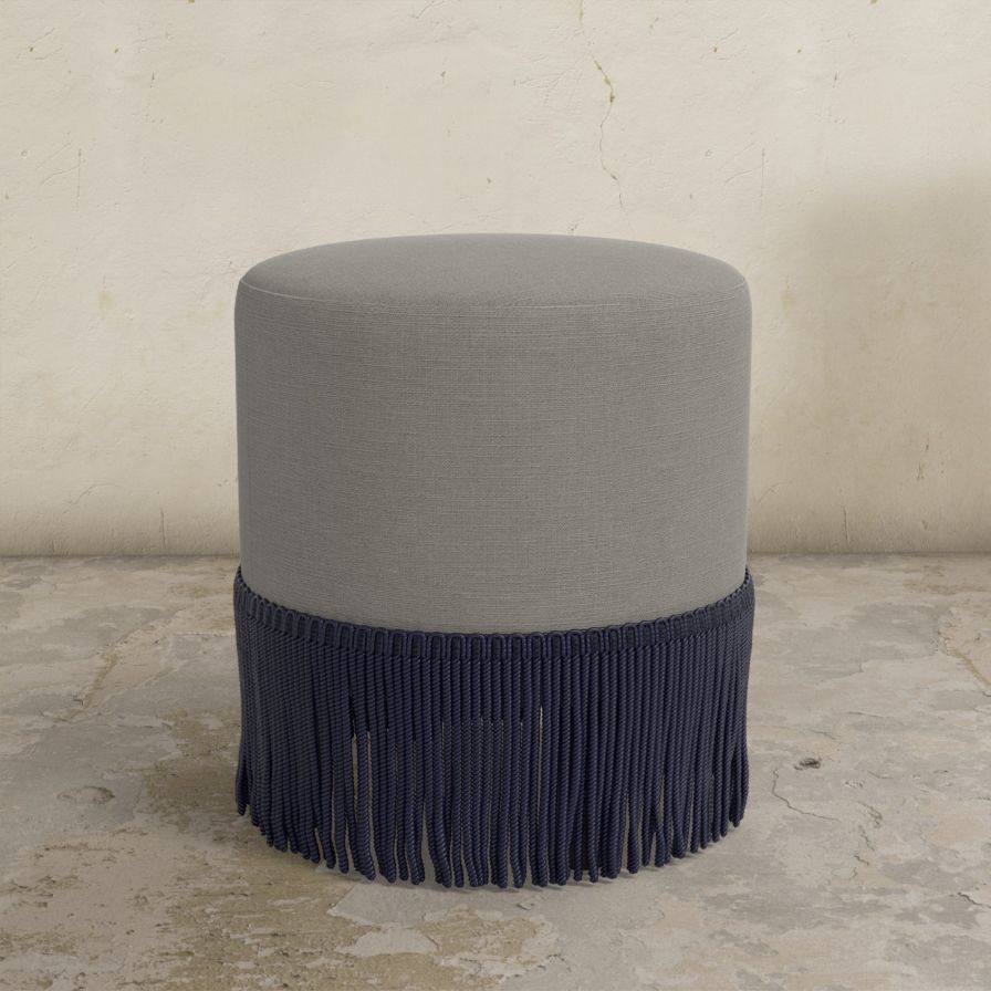 Gray upholstered ottoman with fringe on a concrete floor.