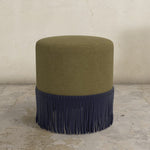 Green upholstered ottoman with  fringe on a concrete floor.