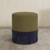 Green upholstered ottoman with  fringe on a concrete floor.