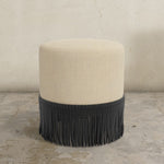 Round upholstered ottoman with beige cushion and black fringe on a concrete floor.