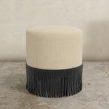 Round upholstered ottoman with beige cushion and black fringe on a concrete floor.
