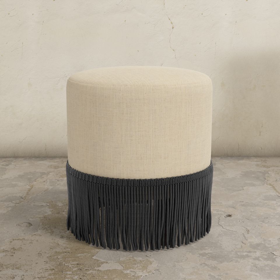Round upholstered ottoman with beige cushion and black fringe on a concrete floor.