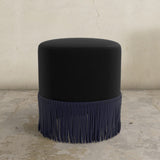 Black upholstered ottoman with fringes on a textured floor and light gray wall background