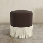 Round ottoman with a dark brown cushion and white fringe on a concrete floor.