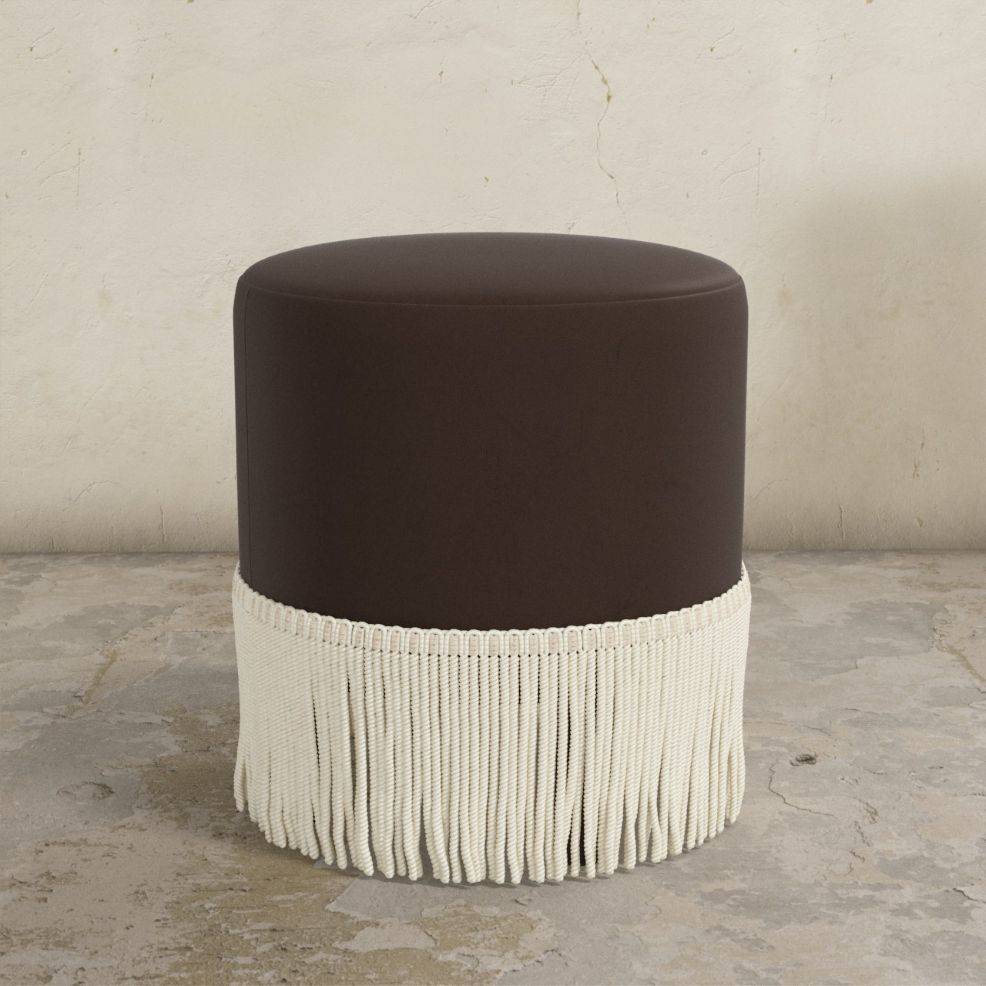 Round ottoman with a dark brown cushion and white fringe on a concrete floor.