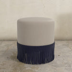 Round ottoman with navy blue fringe on a stone floor.