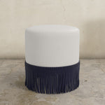 Light grey upholstered ottoman with  fringes on a beige floor.