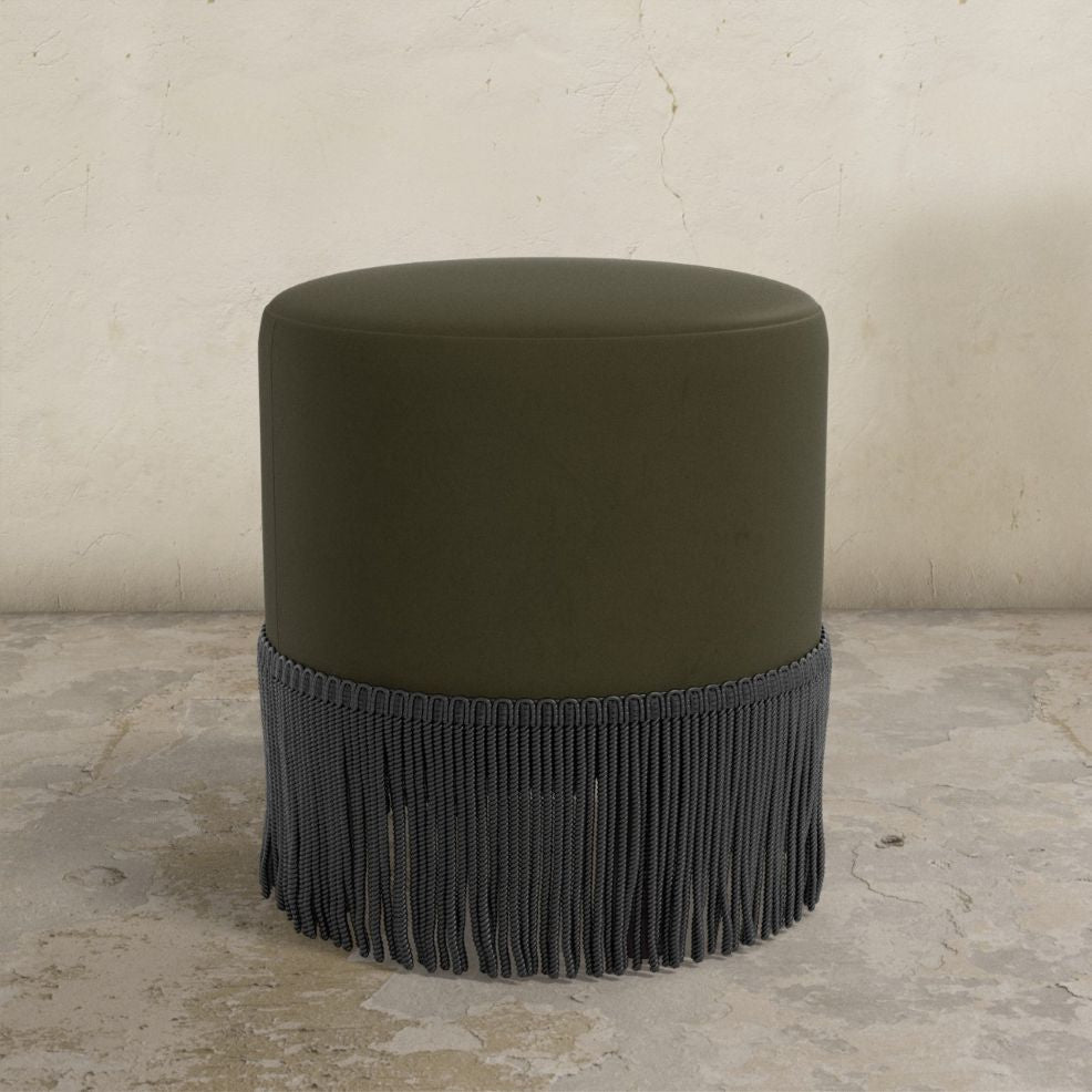 Dark green upholstered ottoman with fringe on a concrete floor and light gray wall background