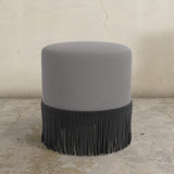 Gray upholstered ottoman with black fringe on a concrete floor.