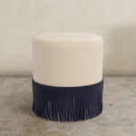 Round upholstered ottoman with black fringe on a textured floor.