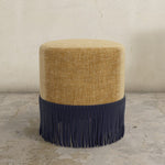 Round upholstered ottoman with beige top and navy fringes on a concrete floor.
