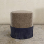 Round upholstered ottoman with brown top and navy fringes on a concrete floor.