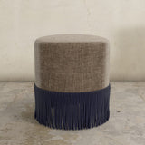 Round upholstered ottoman with brown top and navy fringes on a concrete floor.