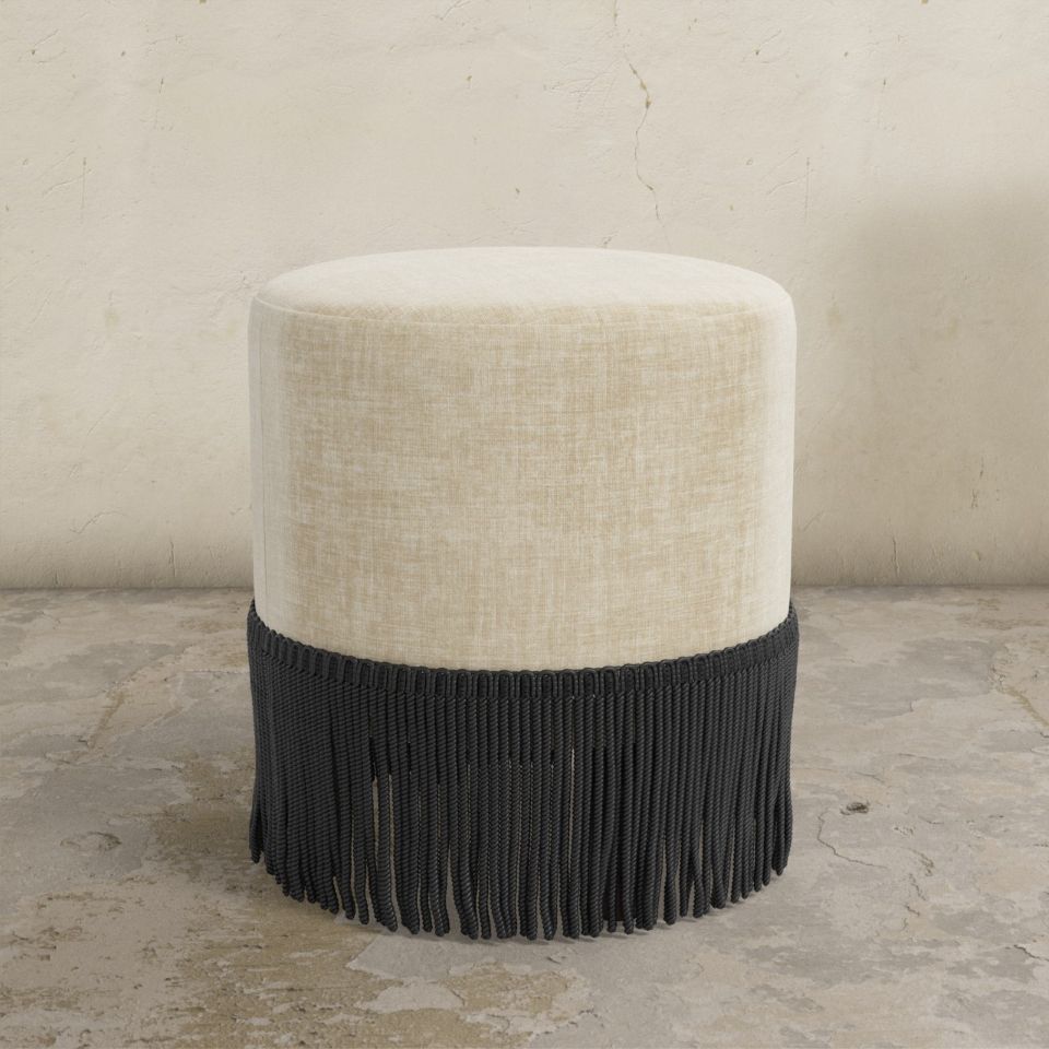 Round upholstered ottoman with beige top and black fringes on a concrete floor.