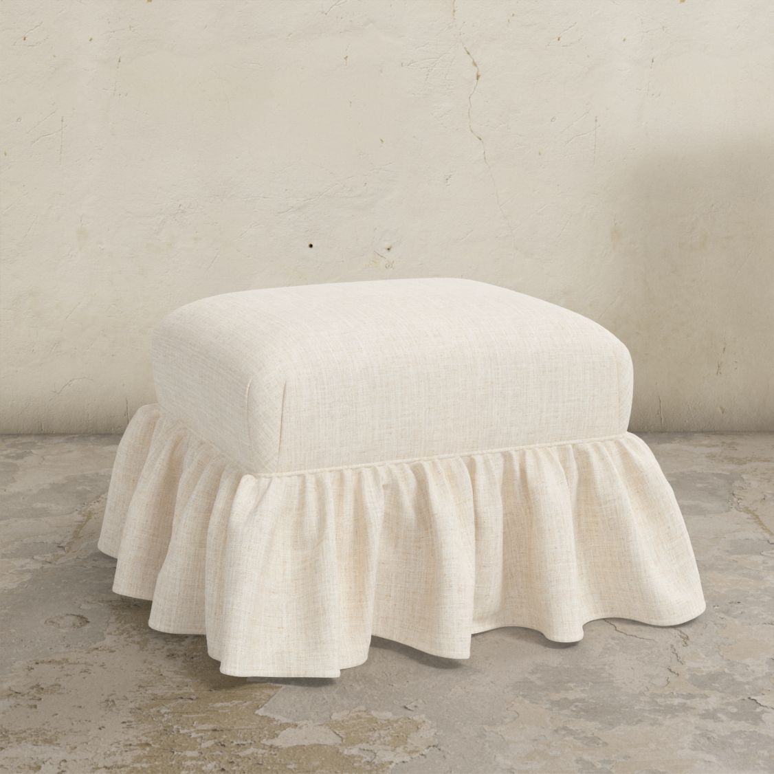 White linen upholstered Edith ottoman with a ruffled skirt on a concrete floor.