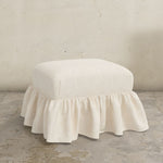 White linen upholstered Edith ottoman with a ruffled skirt on a concrete floor.