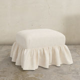 White linen upholstered Edith ottoman with a ruffled skirt on a concrete floor.