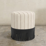 Round upholstered ottoman with striped pattern in neutral tones and fringe on a concrete floor.