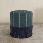 Striped ottoman with fringes on a concrete floor against a beige wall