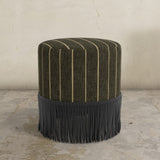 Brown upholstered ottoman with fringe on a concrete floor and light gray wall background