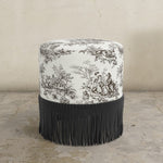 Round upholstered ottoman with patterned top and fringe on a beige background