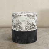 Round upholstered ottoman with patterned top and fringe on a beige background