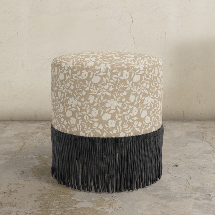Round upholstered ottoman with floral pattern and black fringe on a concrete floor.