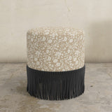 Round upholstered ottoman with floral pattern and black fringe on a concrete floor.