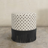 Round upholstered ottoman with patterned top and fringes on a concrete floor.