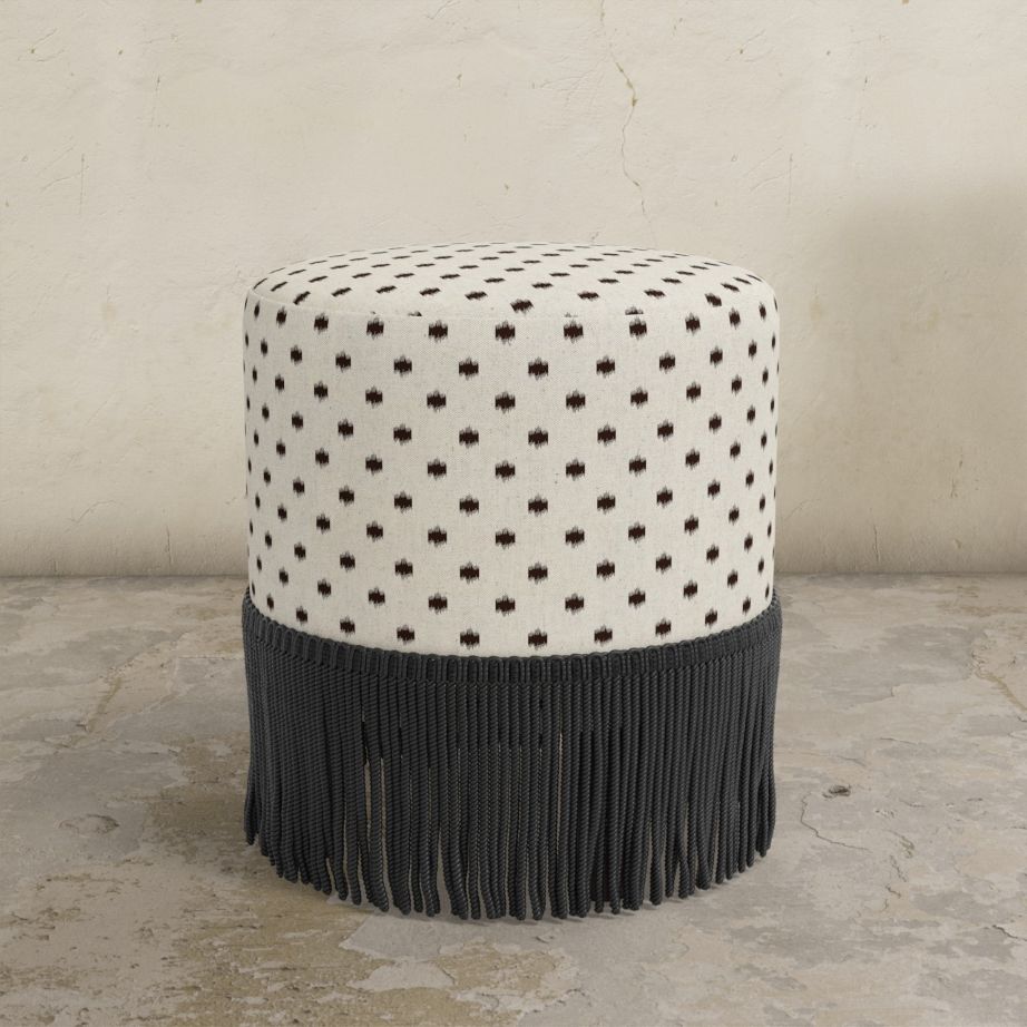 Round upholstered ottoman with patterned top and fringes on a concrete floor.
