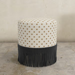 Round upholstered ottoman with patterned top and black fringed bottom on a concrete floor.