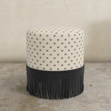 Round upholstered ottoman with patterned top and black fringed bottom on a concrete floor.