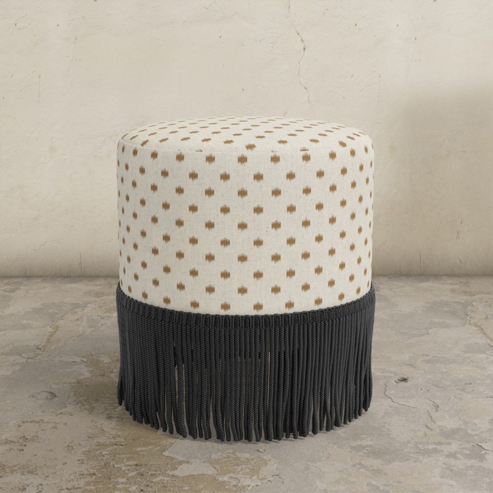 Round upholstered ottoman with patterned top and black fringed bottom on a concrete floor.