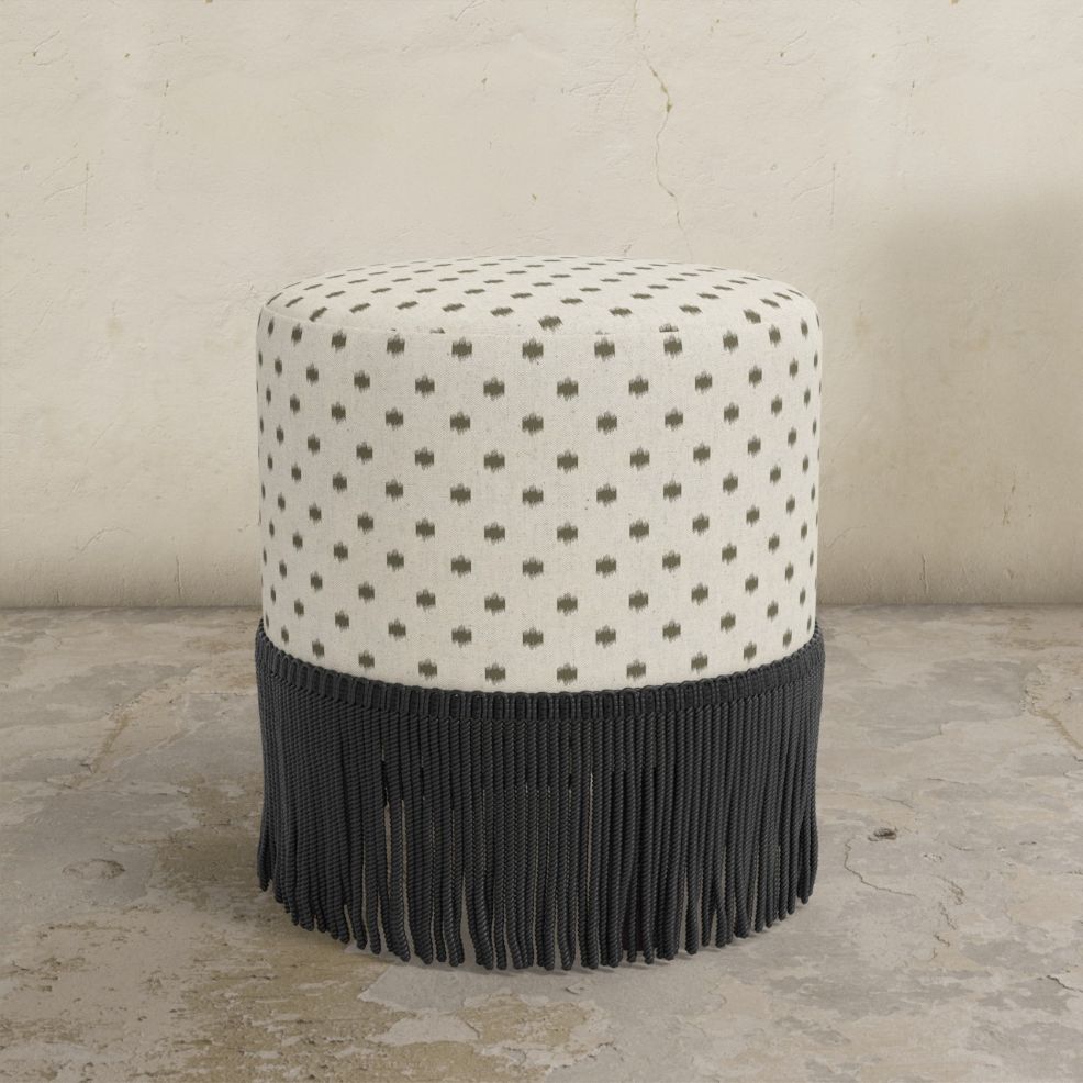 Round upholstered ottoman with patterned top and black fringed bottom on a concrete floor.