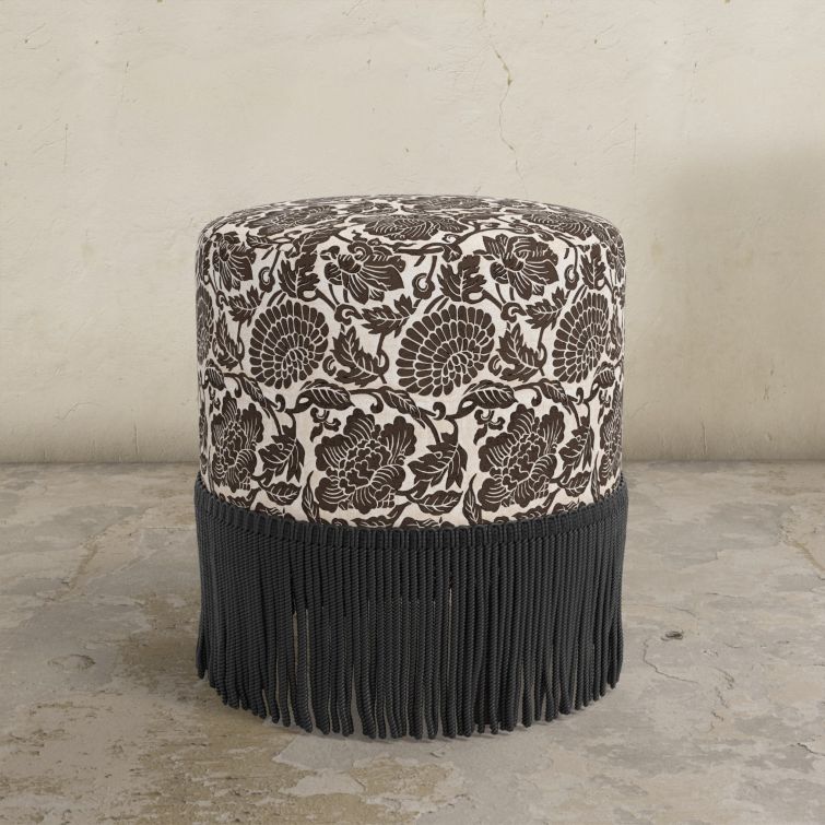 Round upholstered ottoman with floral pattern in chocolate brown and fringe on a concrete floor.